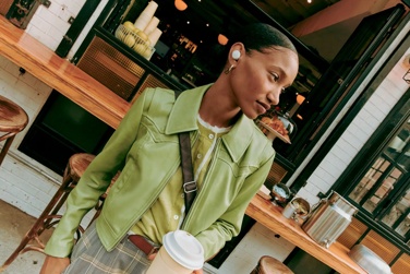 Woman wearing green jacket and QuietComfort Earbuds leaving coffee shop
